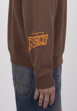 Brązowy rękaw bluzy z pomarańczowym napisem „Legacy Racing” noszony na niebieskich dżinsach, ręka swobodnie oparta wzdłuż boku na jednolitym tle.