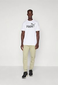 Valkoinen lyhythihainen t-paita, jossa musta Puma-logo, yhdistetty vaaleanvihreisiin kapeisiin juoksuhousuihin ja mustiin urheilukenkiin.