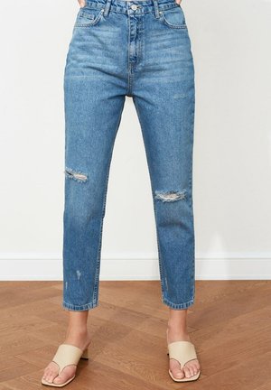 Kobieta ubrana w niebieskie, podniszczone dżinsy z wysokim stanem, z rozdarciami na kolanach, oraz beżowe sandały na obcasie z odkrytymi palcami, stojąca na drewnianej podłodze.