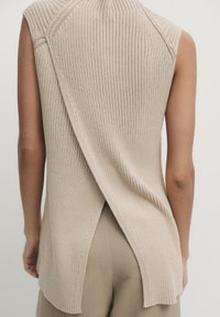 Beżowy, prążkowany, bezrękawnik z krzyżującym się tyłem. Posiada wysoki dekolt i rozcięcia z tyłu, w zestawie z beżowymi spodniami.