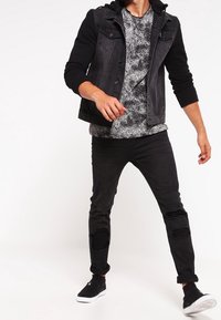 Hombre vistiendo jeans negros rasgados, zapatillas negras, una camiseta gris jaspeada y una chaqueta de mezclilla negra con capucha, de pie en una pose casual.