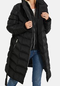 Mujer vestida con chaquetas tipo puffers negras por capas, con cremalleras plateadas y jeans azules, ajustando el cuello de la chaqueta exterior.