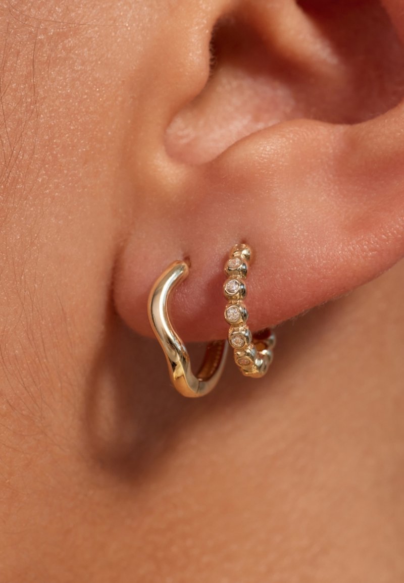 Boucles d'oreilles en or en forme de créoles avec un design ondulé et une deuxième créole ornée de petites pierres claires. Texture et brillance visibles.