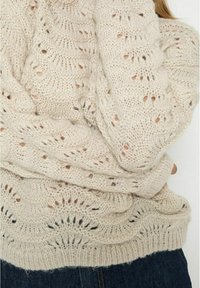 Knit sweater in cream color featuring an openwork design, ribbed hem, and textured fabric. Layered folds create visual depth.