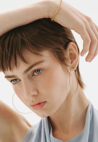 Pendientes de aro dorados, cabello corto y castaño, tela azul suave cayendo, textura de piel natural, diseño minimalista, enfoque en el rostro y los accesorios.