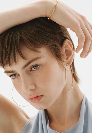 Pendientes de aro dorados, cabello corto y castaño, tela azul suave cayendo, textura de piel natural, diseño minimalista, enfoque en el rostro y los accesorios.