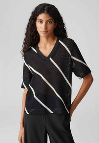 Top corto a maniche corte in rete nera con scollatura a V, caratterizzato da strisce diagonali bianche. Il tessuto è leggero con un motivo a trama aperta.