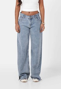 Vaqueros de piernas anchas de color azul claro, hechos de denim, con cintura alta, bolsillos frontales y dobladillos ligeramente deshilachados. Modelo combinado con zapatillas.