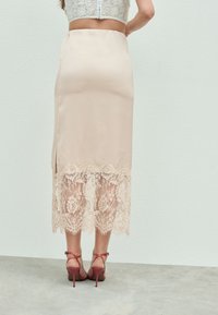 Jupe midi beige avec un corps en satin et une bordure en dentelle délicate à l'ourlet. Présente une fente latérale et est assortie à des talons hauts rouges à lanières.