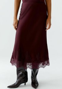 Jupe midi bourgogne avec ourlet en dentelle, présentant des détails en scie intriqués ; associée à des bottes noires brillantes à bout pointu.