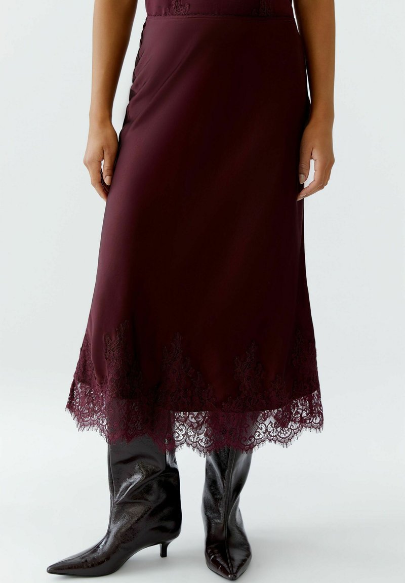 Jupe midi bourgogne avec ourlet en dentelle, présentant des détails en scie intriqués ; associée à des bottes noires brillantes à bout pointu.