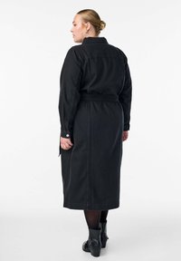 Pikkade mustade vööga paljaste õlgadega mantel, mustad sukkpüksid ja mustad ankle-boots, seljaga valge tausta poole.