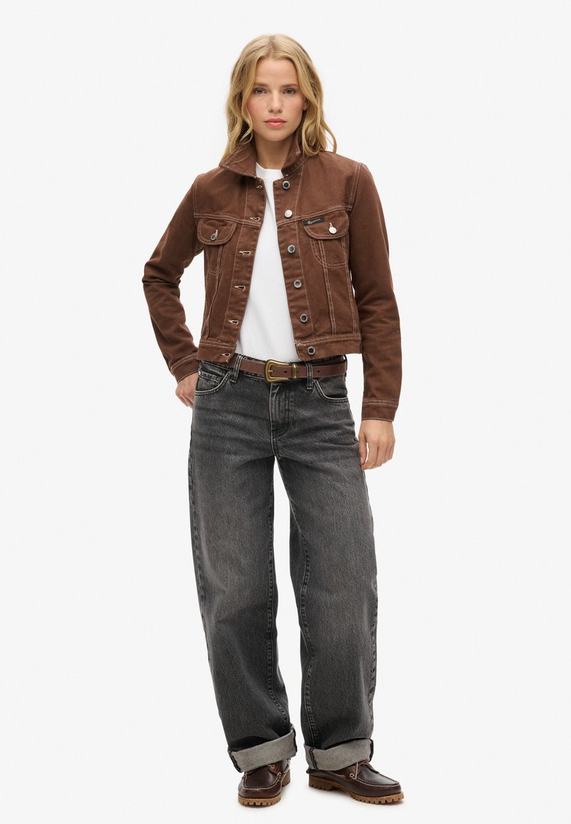 Veste en denim marron avec fermeture à boutons, deux poches poitrine, associée à un jean gris taille haute et coupe ample, ainsi qu'à des chaussures marron foncé.