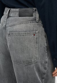 Γκρι τζιν από τζιν με κανονική γραμμή, υφή υφάσματος, και μαύρο δερμάτινο λογότυπο στο πίσω μέρος. Μικρή πολύχρωμη λεπτομέρεια σημαίας στην τσέπη.