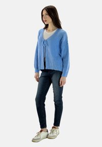 Hellblaue Strickjacke mit V-Ausschnitt, Bindeband vorne und weiten Ärmeln, kombiniert mit dunklen Jeans und weißen Turnschuhen.