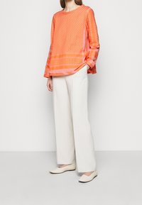 Blusa arancione brillante con motivi geometrici, maniche lunghe e vestibilità rilassata, abbinata a pantaloni bianchi a gamba larga e scarpe basse.