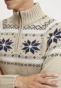 Beige neulepusero, jossa on korkea ribattukraava, sinisiä ja laivastonsinisiä lumihiutalekuvioita sekä lyhyt vetoketju kauluksessa.