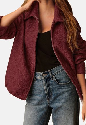 Mujer con una chaqueta burdeos texturizada con cremallera, top negro y jeans azules de cintura alta, con el pelo largo y suelto sobre el hombro.