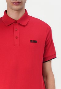 Κόκκινο πόλο μπλουζάκι από μαλακό ύφασμα, με γιακά, τριών κουμπιών και μικρό μαύρο λογότυπο στο αριστερό στήθος. Μαύρες λεπτομέρειες στα μανίκια.