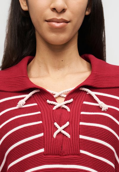 Γυναίκα που φοράει πλεκτό τοπ με κόκκινες και λευκές ρίγες, με λευκές λεπτομέρειες κορδονιού στη λαιμόκοψη, που αποκαλύπτει τον λαιμό και το κάτω μέρος του προσώπου.