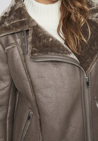Giacca in shearling grigia con colletto in morbido marrone, dotata di chiusura con zip e pelle testurizzata. Include una tasca laterale con zip.