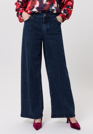 Donkere denim jeans met wijde pijpen, contrasterend lichte stiksels, een hoge taille en voorzakken, gecombineerd met een levendige rode blouse met patroon en roze hakken.