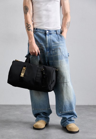Black duffel bag with gold hardware and logo, made of textured fabric. Worn with light blue, distressed wide-leg jeans and beige clogs.