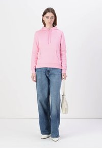 Hoodie en coton rose clair avec une poche avant et un capuchon à cordon, assorti à un jean bleu à jambes larges et des chaussures blanches, tenant un sac beige.