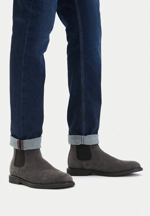 Osoba nosící ohrnuté tmavě modré džíny a šedé semišové Chelsea boty na bílém pozadí.