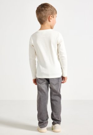 Jonge jongen die met zijn rug naar de kijker staat, gekleed in een wit shirt met lange mouwen, grijze spijkerbroek en witte sneakers, tegen een effen achtergrond.