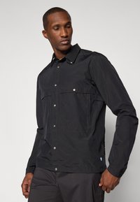 Black jacket made of lightweight fabric with a button-up front, two chest pockets, and adjustable cuffs. Features a small logo on the side.