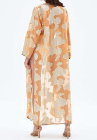 Žena oblečená v dlouhém, průhledném oranžovo-stříbrném hábitu s bočním rozparkem, doplněná nahými lodičkami na vysokém podpatku, zobrazená zezadu.