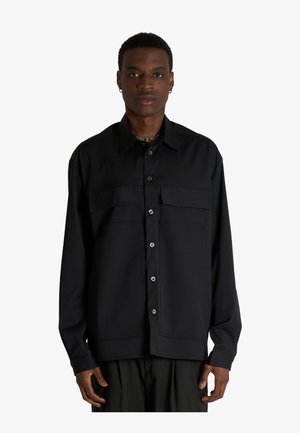 Uomo con capelli corti che indossa una camicia nera a maniche lunghe con bottoni e tasche sul petto, in piedi davanti a uno sfondo bianco uniforme.