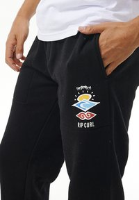 Sorte sweatpants lavet af blødt stof med et logo design. Logoet inkluderer en sol, bølge grafik og teksten "RIP CURL."