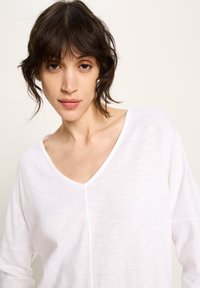 Junge Frau mit kurzem dunklem Haar, die ein weißes Top mit V-Ausschnitt trägt und direkt in die Kamera schaut, vor einem schlichten hellen Hintergrund.