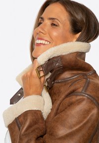 Braune Lammfelljacke mit strukturierter Oberfläche, beigem Fleecekragen und verstellbarem Messingzubehör am Kragen.