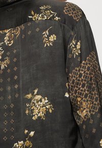 Camicia in tessuto nero con motivi floreali in oro e beige, arricchita da dettagli geometrici; texture liscia e design strutturato con un logo discreto.