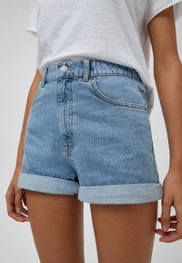 Person i ljusblå uppvikt denimshorts och en vit t-shirt, sedd från midjan till halva låret mot en neutral bakgrund.