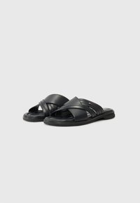 Sandales à glissière en cuir noir avec des brides croisées, une semelle plate et des détails texturés subtils sur les brides.