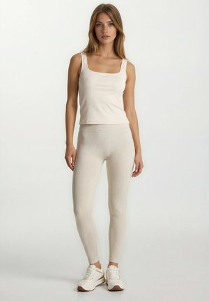 Nainen pukeutunut kermaiseen ribbineulottuun tankkitoppiin ja siihen sopiviin legginsseihin, seisoo suorassa, jalassa valkoiset lenkkarit, neutraali tausta.