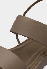 Καφέ δερμάτινο σανδάλι με φαρδιές μπροστινές και αστραγάλου λουράκια, με εμφανείς ραφές κατά μήκος της άκρης της σόλας.