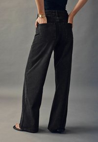 Černé široké džíny vyrobené z denimové látky s texturovaným povrchem. Obsahují dvě zadní kapsy a dekorativní prošívání podél bočních švů.