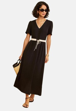 Donna con abito midi nero a scollo a V con cintura bianca, occhiali da sole neri, tiene una borsa di paglia e indossa sandali neri con cinturini.