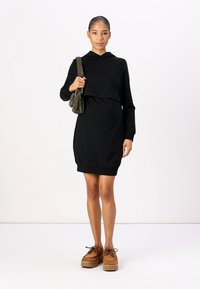 Robe à capuche noire avec un design cropped, des poignets côtelés et un ourlet arrondi. Le modèle tient un petit sac olive et porte des chaussures à plateforme beiges.