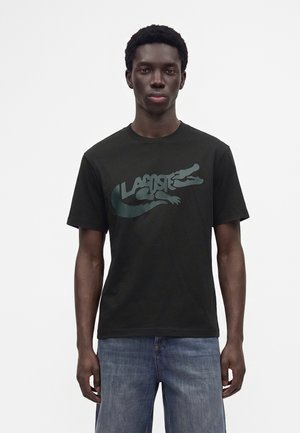 Jeune homme portant un t-shirt noir Lacoste avec un grand logo crocodile et un jean bleu, debout devant un fond clair uni.