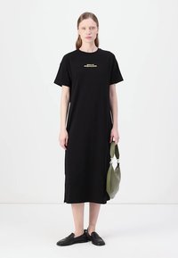 Fekete pamut pólóruha rövid ujjakkal, térdhez érő hosszúságú, arany színű grafikai nyomattal a mellrészen. A modell egy zöld táskát tart.