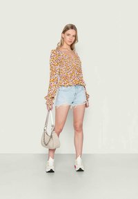 Blusa floral amarela com mangas em babados, combinada com calções de ganga desbotados. Acompanhada por uma mala de ombro cinza claro e ténis brancos desportivos.