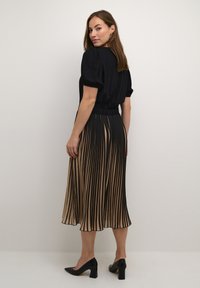 Juodas viršus su pūstomis rankovėmis, elastinga juosta ir klostuota smėlio spalvos sijonas su juodomis juostelėmis. Nešiojama su juodais kulniukais.