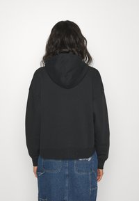 Sudadera negra corta con capucha, presenta un diseño sencillo y tela texturizada. La parte trasera muestra costuras en los hombros y un ajuste holgado.