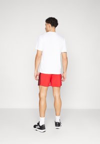 White short-sleeve t-shirt paired with red shorts featuring black side stripes. The model wears black athletic shoes and white socks.